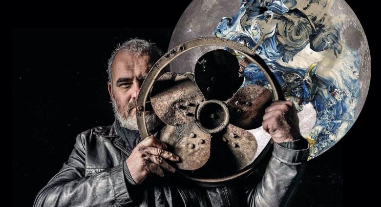 artist standing with metal fan in front of moon painting/ sculpture