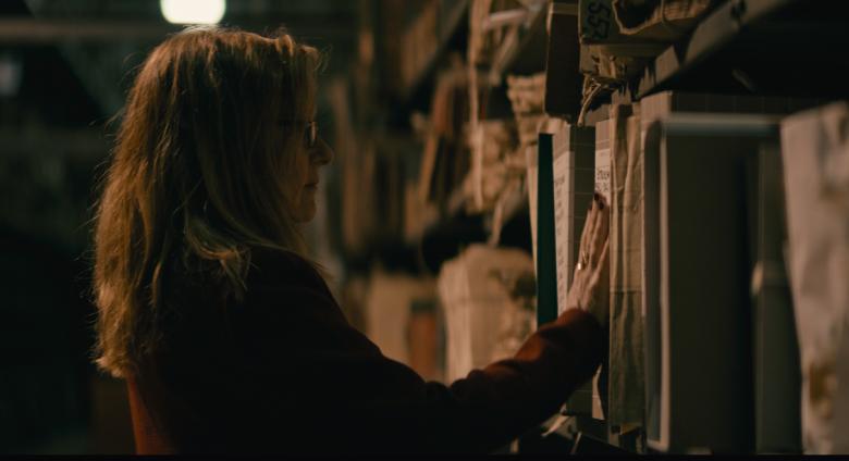 Lynda is seen in dark room, looking at shelf packed with documents. 