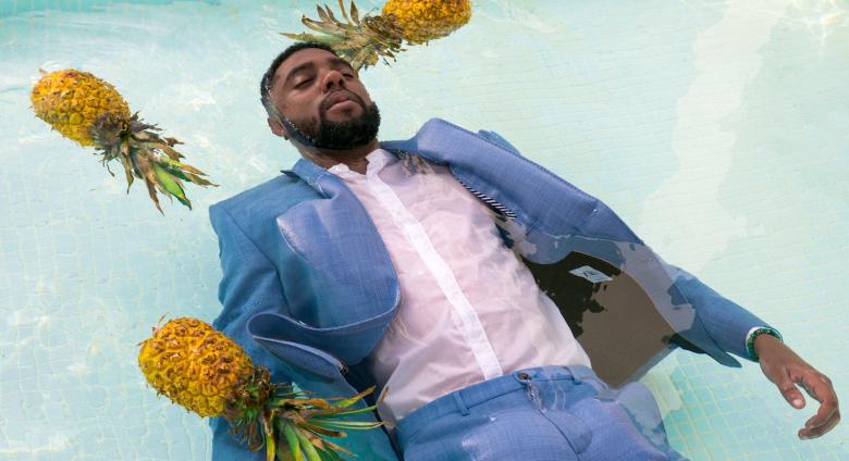 A man with dark skin, wearing a suit floats in a pool amongst three pineapples. 