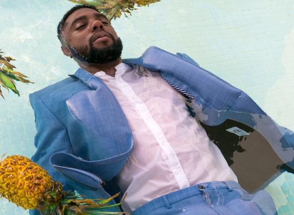 A man with dark skin, wearing a suit floats in a pool amongst three pineapples. 